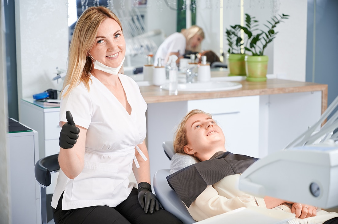 female stomatologist showing approval gesture and smiling while woman lying in dental chair